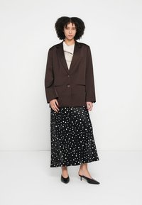Brown blazer with a single button and two pockets, paired with a black satin skirt featuring white polka dots. Sharp pointed black heels.