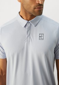 Lyseblå polo med tekstureret overflade, knapkrave og sort Nike-logo på venstre bryst. Korte ærmer, åndbart stof.