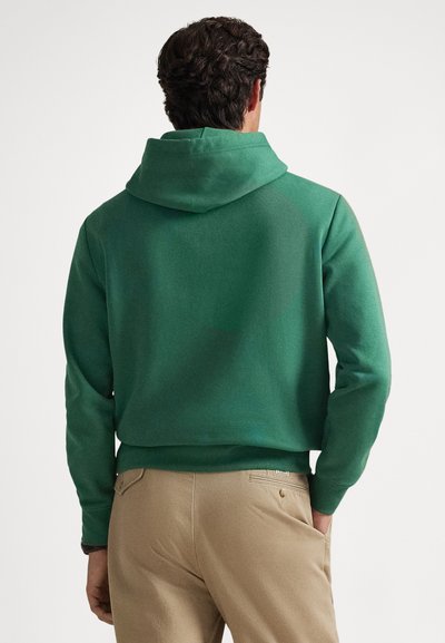 Vyras su tamsiais garbanotais plaukais, vilkintis žalią džemperį su gobtuvu ir smėlio spalvos kelnes, stovintis su viena ranka kišenėje, atsisukęs nuo žiūrovo.