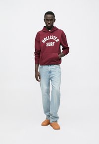 Bordo majica s kapuljačom s natpisom "HOLLISTER SURF", svijetloplave traperice i smeđa obuća. Majica s kapuljačom ima džep sprijeda i podesivu kapuljaču s vezicama.