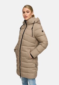 Young woman with blonde hair wearing a beige padded long coat with hood and black zipper, hands in pockets, against a plain background.