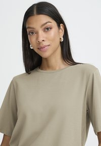 Kvinde med langt mørkt hår iført sølvfarvede øreringe i ring og en beige kortærmet skjorte, der roligt kigger på kameraet mod en hvid baggrund.