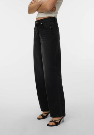 Personne portant un jean noir taille haute à jambes larges et des sandales à talons noires à lanières, debout les bras croisés devant un fond uni.