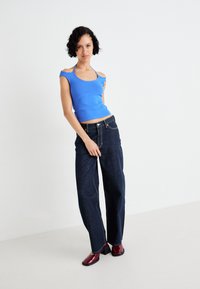 Modra prilegajoča se majica s izrezanimi rameni, v kombinaciji s temnimi širokimi denim hlačami. Model nosi rdeče čevlje na blok peto in stoji na svetli podlagi.