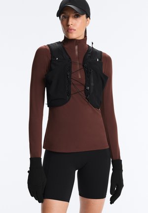 Frau trägt schwarze Mütze, schwarze taktische Weste mit vorderen Schnüren, braunes langärmeliges Oberteil, schwarze Handschuhe und schwarze Radlerhosen, steht vor weißem Hintergrund.