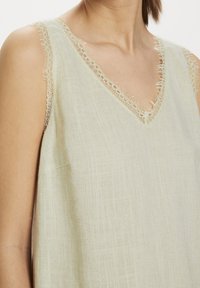 Top in lino beige chiaro con scollo a V e delicata rifinitura in pizzo attorno al collo e alle aperture delle maniche. Tessuto morbido e leggermente strutturato.