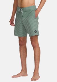 Bañador verde para hombre con rayas blancas horizontales, cintura elástica, cierre con cordón y un parche de logo negro en la pierna inferior.