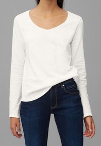 Vrouw draagt een effen wit shirt met lange mouwen en een V-hals, dat lichtjes in een donkerblauwe spijkerbroek is gestopt, staand tegen een grijze achtergrond.