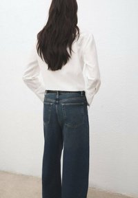 Camisa blanca de manga larga combinada con pantalones vaqueros de pierna ancha de talle alto en azul oscuro, con bolsillos traseros y textura suave.