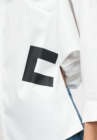 Λευκό πουκάμισο με μεγάλο μαύρο κεφαλαίο ελληνικό γράμμα Σίγμα στο κάτω μέρος της πλάτης, φορεμένο με μπλε τζιν, το χέρι λυγισμένο πίσω από την πλάτη.