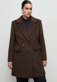 Cappotto doppiopetto marrone con grandi rever, due tasche laterali e bottoni arrotondati; il tessuto strutturato conferisce un look definito.