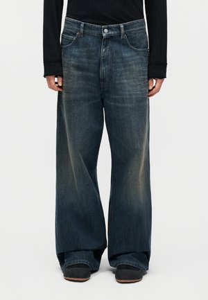 Άτομο που φοράει άνετο, φαρδύ παντελόνι τζιν σε ξεθωριασμένο σκούρο μπλε χρώμα, με μαύρο μακρυμάνικο μπλουζάκι και μαύρα παπούτσια, στέκεται μπροστά σε ένα απλό ανοιχτόχρωμο φόντο.