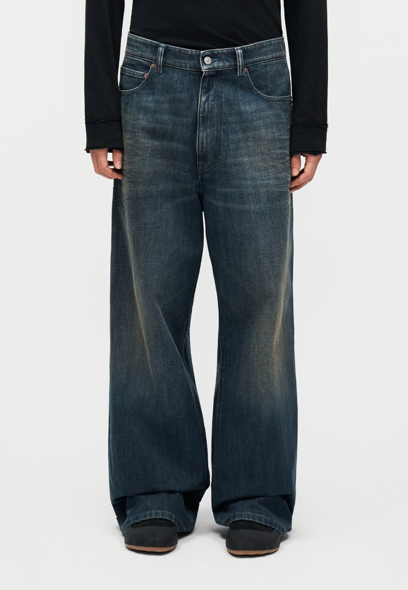 Personne portant un jean bleu foncé délavé, ample et à jambes larges, un haut à manches longues noir et des chaussures noires, debout devant un fond clair uni.