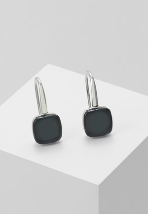Boucles d'oreilles pendantes en argent avec des pierres carrées noires, présentées sur un socle blanc sur un fond gris.