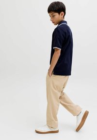 Garçon en polo bleu marine et pantalon beige marchant les mains dans les poches sur un fond clair uni.