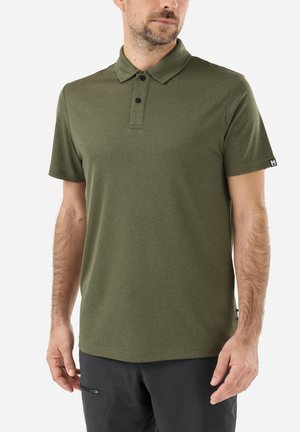 Homme portant un polo vert olive à manches courtes avec des boutons noirs et un pantalon noir, debout devant un fond uni.