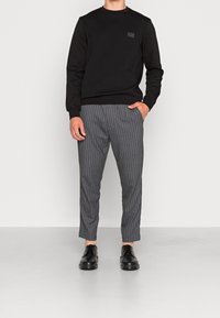 Felpa nera con colletto rotondo e una piccola patch con logo, abbinata a pantaloni grigi a righe e scarpe nere con lacci.