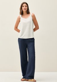 Haut sans manches blanc avec des bretelles décoratives, associé à un pantalon bleu marine ample. Des sandales beiges avec des détails tissés complètent la tenue.