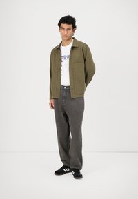 Olive green jacket with buttons, white t-shirt featuring purple graphic, dark gray pants, and black sneakers with white stripes.