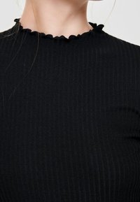 Haut noir côtelé avec un col montant et un bord festonné. Le tissu présente une finition texturée, mettant en valeur sa coupe ajustée.