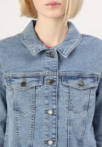 Jasnoniebieska kurtka dżinsowa o klasycznym fasonie, z kołnierzykiem w kształcie punktu, zapięciem na guziki z przodu oraz dwiema kieszeniami na piersi z metalowymi akcentami.