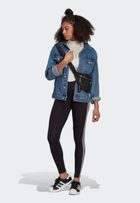 Giacca di jeans con chiusura a bottoni, dolcevita bianco, leggings neri con strisce bianche e sneakers nere con dettagli bianchi.