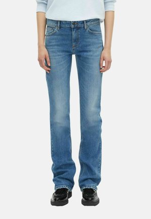 Persoon in lichtblauwe recht-toe-rechtaan jeans en zwarte loafers, met ontspannen handen langs de zijden tegen een witte achtergrond.