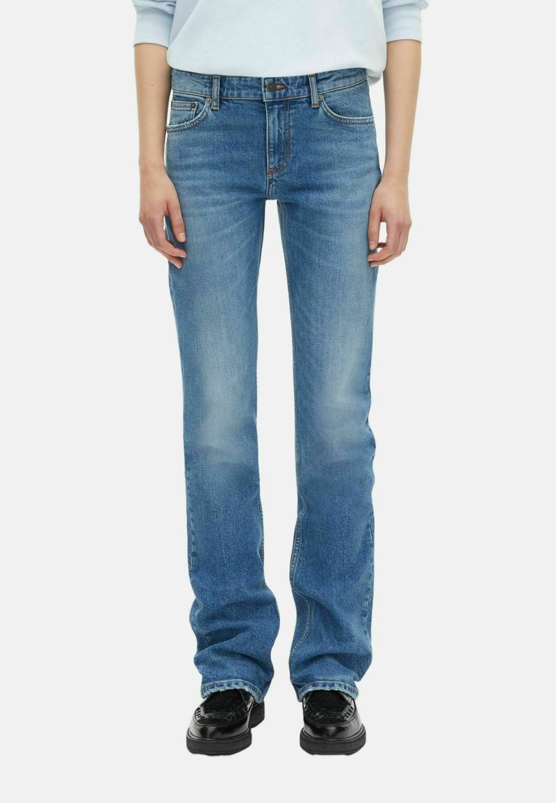 Personne portant un jean droit bleu clair et des mocassins noirs, les mains détendues sur les côtés, sur un fond blanc.