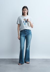 Ung kvinne iført grå t-skjorte med motiv av teddybjørn, høytlivs blå jeans, svarte sandaler, og holder en hvit skulderveske.