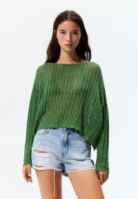 Grüner, gestrickter Pullover mit einem lockeren, offenen Webdesign, kombiniert mit zerrissenen Denim-Shorts. Helle Farbe und strukturierter Stoff.