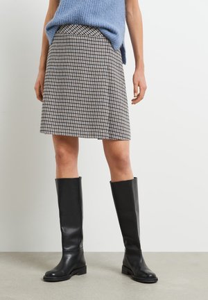 Femme portant des bottes en cuir noires jusqu'aux genoux, une jupe à carreaux longueur genou et un haut en tricot bleu sans manches, debout sur un sol gris.