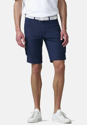 Man draagt marineblauwe cargoshorts, witte riem, witte sneakers en een wit shirt, staand tegen een lichte achtergrond.