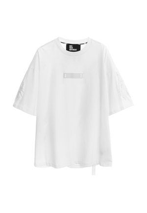 Camiseta blanca oversize de manga corta con cuello redondo, con sutiles diseños de alas bordadas en las mangas y un pequeño parche rectangular en el pecho.