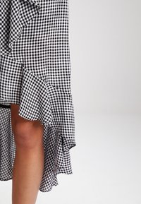 Jupe à motif vichy noir et blanc avec un ourlet à volants, fabriquée en tissu léger, dotée d'une coupe asymétrique qui dévoile la jambe.