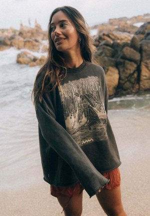 Szara, oversize bluza z nadrukiem graficznym przedstawiającym nadmorski krajobraz, z długimi rękawami i luźnym krojem. Noszona z wzorzystymi szortami.