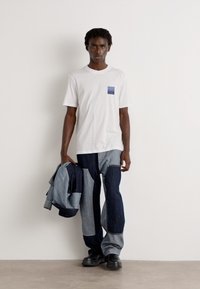 White t-shirt with a blue pocket, paired with patchwork denim pants in varying shades of blue. Holding a denim jacket; black boots.