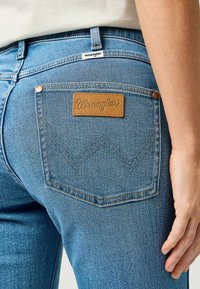 Jeans de mezclilla de color azul claro con un parche de cuero marrón claro que lleva el logotipo "Wrangler" en relieve, bolsillos traseros en ángulo y un diseño clásico de cinco bolsillos.