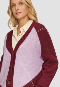 Strickjacke mit einem burgunderfarbenen und lavendelfarbenen Rautenmuster, V-Ausschnitt, Knopfleiste und strukturiertem Stoff. Akzentuiert mit braunen Knöpfen.