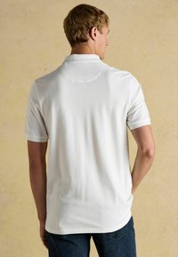 Bílá polo košile s krátkým rukávem, texturovaná tkanina, zaoblený límeček a přiléhavý střih, nošena s modrými džíny na neutrálním pozadí.