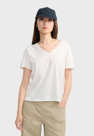 Femme portant un t-shirt blanc à col en V et manches courtes, un pantalon beige avec des poches, et une casquette de baseball bleu foncé, debout avec une main dans la poche.
