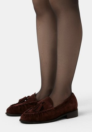 Jambes dans des collants noirs transparents portant des mocassins en daim marron foncé avec des flamboyants, se tenant sur une surface blanche.