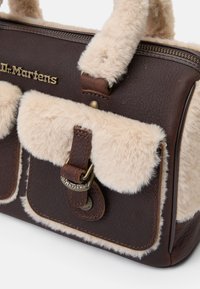 Barna bőr Dr. Martens kézitáska bézs műszőrme díszítéssel, elülső csattal záródik, és puha fogantyúval rendelkezik, fehér háttér előtt.