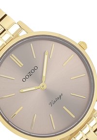 Montre en acier inoxydable doré avec un boîtier rond, un cadran texturé gris clair, des index horaires en or jaune et un bracelet en maillons métalliques.