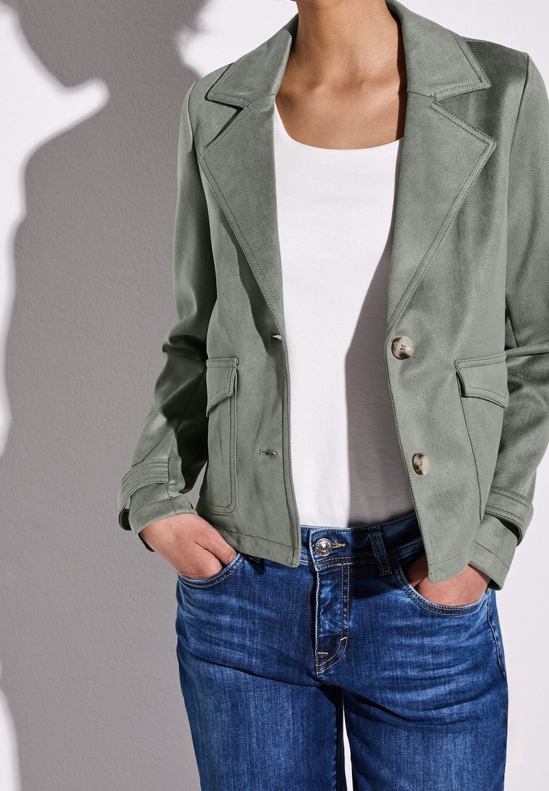 Personne portant un blazer vert clair sur une chemise blanche, les mains dans les poches d'un jean bleu, sur un fond gris.