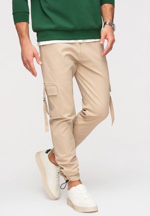 Mężczyzna w beżowych spodniach bojówkach z bocznymi kieszeniami, białych trampkach, zielonej bluzie i srebrnym zegarku na nadgarstku, stoi ze skrzyżowanymi nogami.