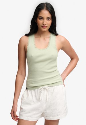 Ung kvinde med langt mørkt hår iført en lysegrøn ribbet tanktop og hvide broderede shorts med snørelukning, står med den ene hånd på hoften.