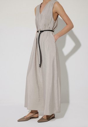 Ujjatlan világos bézs maxi ruha mély V-nyakkivágással, derékban vékony fekete övvel szűkítve, taupe hegyes orrú pántos lapos cipővel viselve.