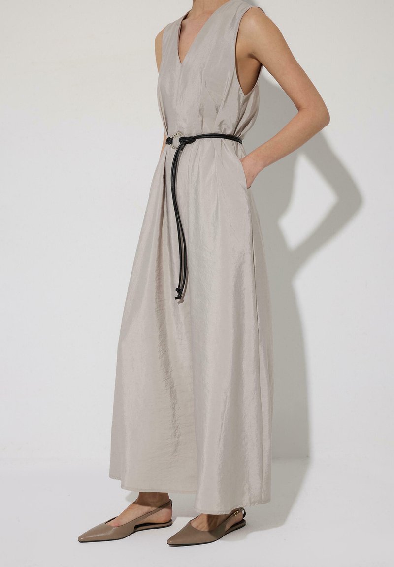 Robe maxi beige clair sans manches avec un décolleté en V profond, taille cintrée par une fine ceinture noire, portée avec des ballerines slingback taupe à bout pointu.