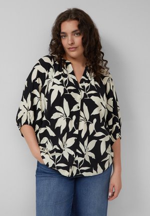 Frau mit lockigem Haar, die ein schwarzes Hemd mit Knöpfen und großem weißem Blumenmuster sowie blaue Jeans trägt, steht vor einem grauen Hintergrund.