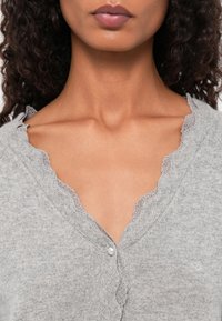 Cardigan cinza feito de material de malha suave, apresentando um decote de renda com estilo ondulado e um fecho com botões, com uma textura delicada por todo o lado.